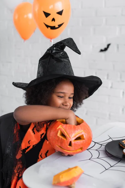 Cheerful african american girl in witch halloween costume holding caved pumpkin — Stock Photo