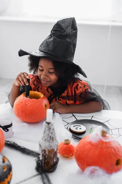 Lächelndes afrikanisch-amerikanisches Mädchen im Hexen-Halloween-Kostüm, das Kürbis schnitzt — Stockfoto