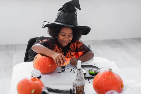 Cheerful african american girl in witch halloween costume near pumpkins and cookies on table — Stock Photo