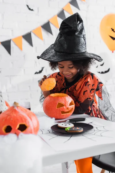 Menina americana africana feliz no traje bruxa halloween olhando para abóbora cavada — Fotografia de Stock