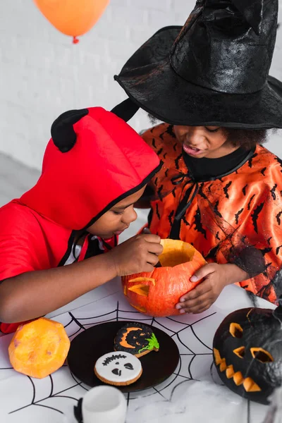 Crianças africanas americanas em trajes de Halloween esculpindo abóbora perto de biscoitos — Fotografia de Stock