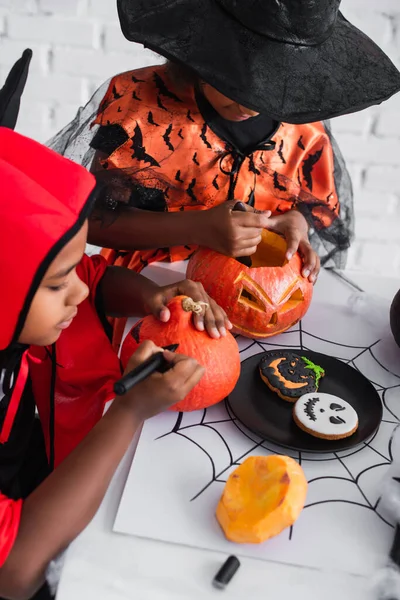 African american kids in halloween costumes carving and drawing on pumpkins near cookies — Stock Photo