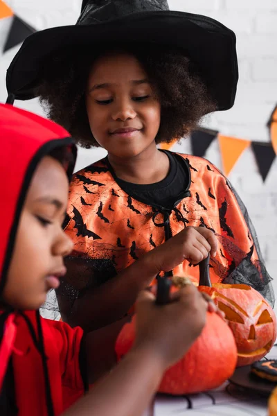 Sorridente ragazza africana americana in costume di Halloween guardando il fratello sfocato che disegna sulla zucca — Foto stock