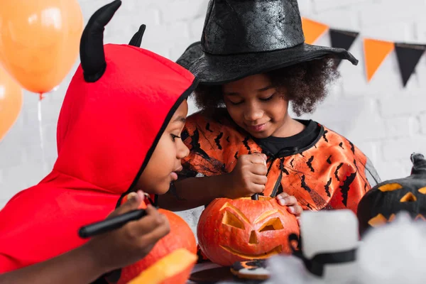 Africano americano chico en halloween traje mirando hermana tallado calabaza - foto de stock