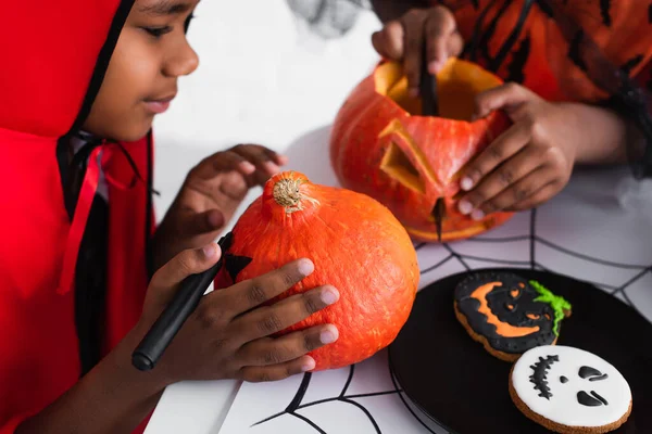 African american boy in halloween costume looking at sister carving pumpkin near cookies — Stock Photo