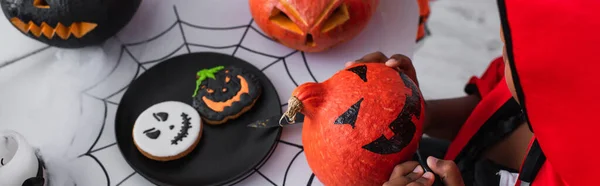 Vista recortada de niño afroamericano en traje de Halloween dibujo en calabaza, pancarta - foto de stock