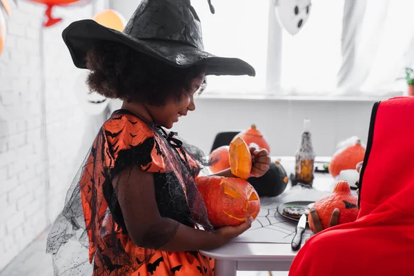 Happy african american girl in pointed hat holding carved pumpkin near brother — Stock Photo