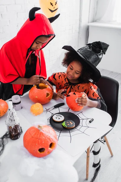 Niños afroamericanos felices en disfraces de Halloween tallando calabazas cerca de galletas en la mesa - foto de stock