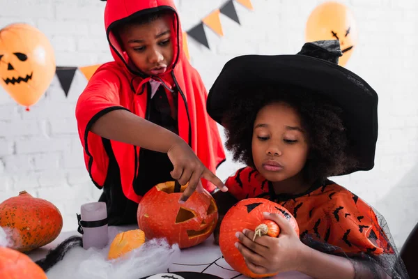 African american boy pointing at carved pumpkin near sister in witch costume — Stock Photo