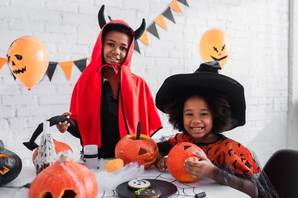 Crianças africanas felizes americanas em trajes de Halloween perto de abóboras esculpidas e biscoitos na mesa — Fotografia de Stock