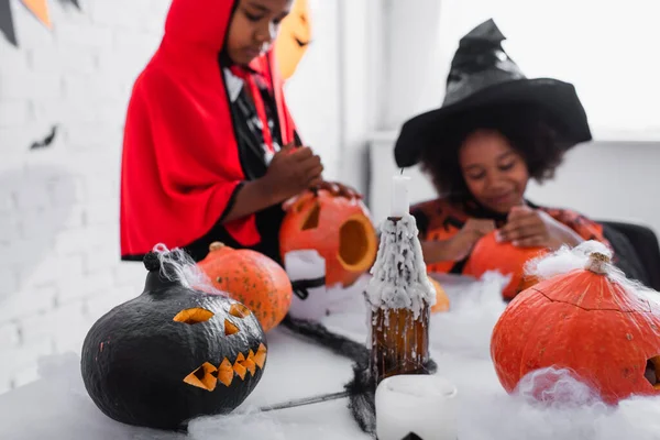 Calabazas talladas y velas cerca de niños afroamericanos borrosos en trajes de halloween - foto de stock