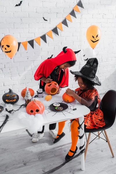 Afrikanisch-amerikanische Kinder in Halloween-Kostümen schnitzen Kürbisse in der Nähe von Plätzchen auf dem Tisch — Stockfoto