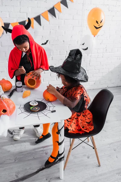 Bambini africani americani in costumi di Halloween intaglio zucche vicino biscotti fatti in casa sul tavolo — Foto stock