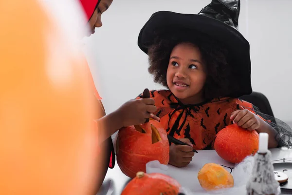 Feliz afroamericano chica en traje de bruja mirando hermano tallado calabaza - foto de stock