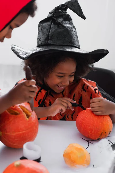 Offuscata africano americano ragazzo intaglio mentre felice sorella in appuntito cappello disegno su zucca — Foto stock