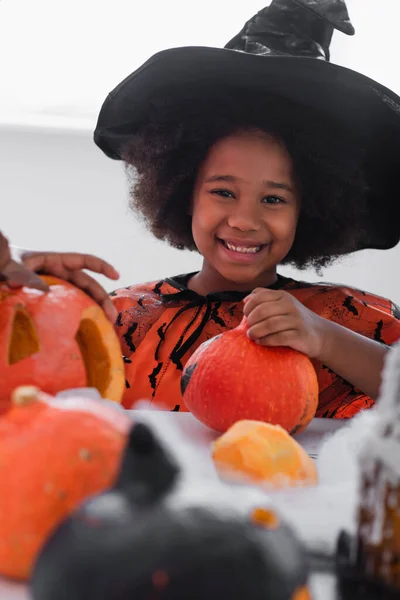 Happy african american girl in witch costume near pumpkins and blurred brother — Stock Photo