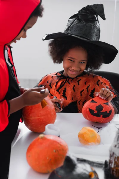 Happy african american girl in witch costume showing drawing on pumpkin near blurred brother — Stock Photo