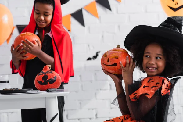 Happy african american girl in witch costume showing pumpkin with drawing near blurred brother — Stock Photo