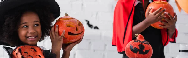 Happy african american girl in witch costume showing pumpkin with drawing near blurred brother, banner — Stock Photo