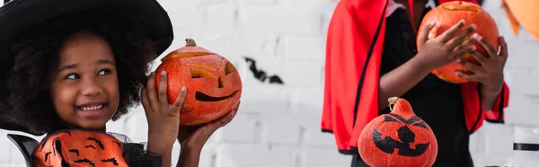 Feliz afroamericano chica en traje de bruja mostrando calabaza con dibujo cerca borrosa hermano, bandera - foto de stock