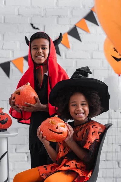 Happy african american kids in halloween costumes holding carved pumpkins at home — Stock Photo