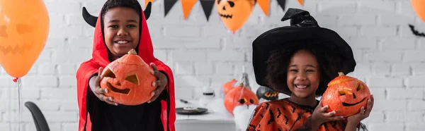 Crianças americanas africanas alegres em trajes de Halloween segurando abóboras esculpidas em casa, bandeira — Fotografia de Stock