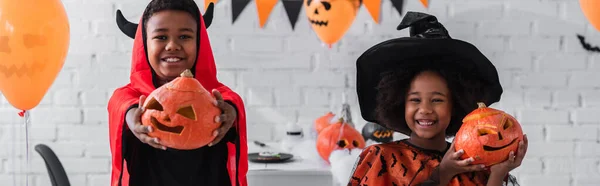 Cheerful african american kids in halloween costumes holding carved pumpkins at home, banner — Stock Photo