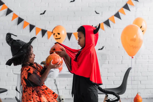 Vista lateral de crianças americanas africanas em trajes de Halloween segurando abóboras esculpidas em casa — Fotografia de Stock