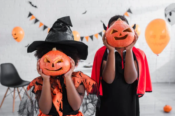 African american kids in halloween costumes covering faces with carved pumpkins at home — Stock Photo