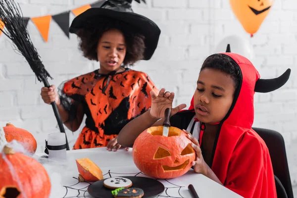 Africano americano ragazzo in halloween costume scultura zucca vicino sfocata sorella con scopa — Foto stock