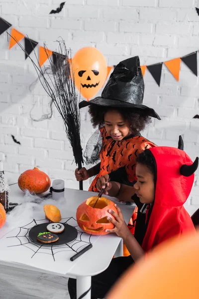 African american boy in halloween costume carving pumpkin near sister with broom — Stock Photo