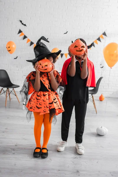 Longitud completa de los niños afroamericanos en disfraces de Halloween cubriendo caras con calabazas talladas en casa - foto de stock