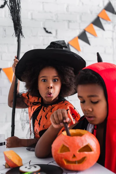 Offuscata africano americano ragazzo in halloween costume scultura zucca vicino spettrale sorella con scopa — Foto stock