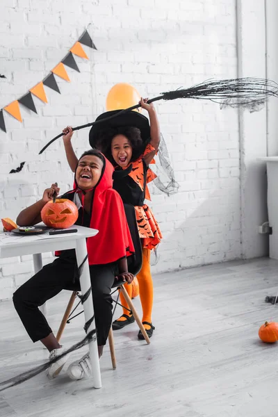 Щасливий афроамериканський хлопчик у костюмі Хеллоуїна різьблення гарбуза біля веселої сестри з мітлою — стокове фото