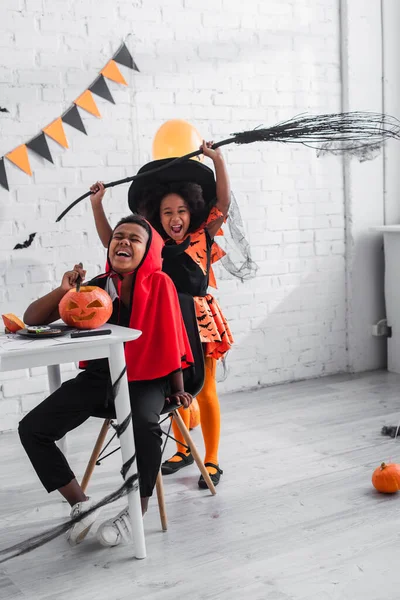 Felice ragazzo africano americano in costume di Halloween intaglio zucca vicino sorella allegra con scopa — Foto stock