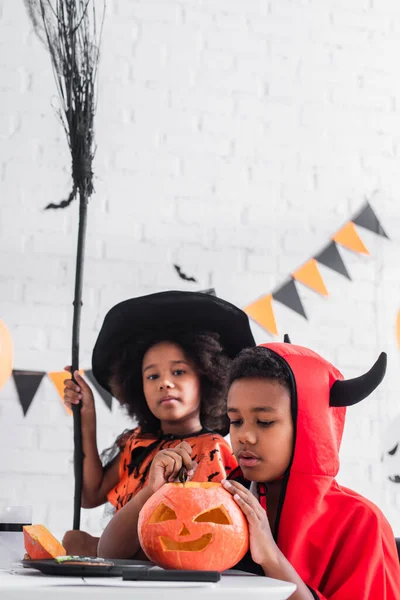 Blurred african american boy carving pumpkin near sister in witch halloween costume with broom — Stock Photo