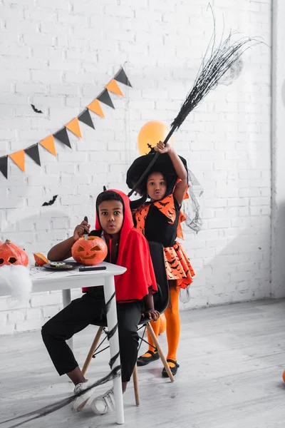 Афро-американський хлопчик у костюмі Хеллоуїна різьблення гарбуза біля сумної сестри з мітлою — стокове фото