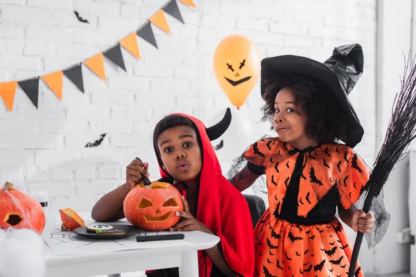 Spooky african american boy in halloween costume carving pumpkin near sister with broom — Stock Photo