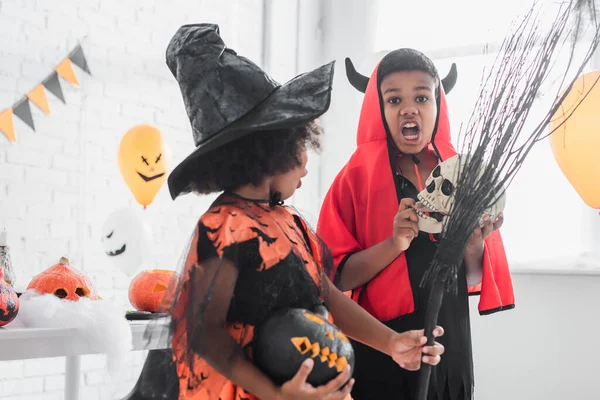 Assustador afro-americano menino em halloween traje segurando crânio perto irmã com vassoura e abóbora esculpida — Fotografia de Stock