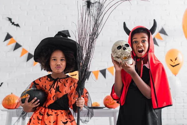 Creepy african american boy in halloween costume holding skull near spooky sister with broom and carved pumpkin — Stock Photo