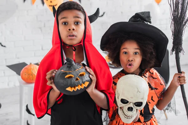 Spooky african american children in halloween costumes holding skull, carved pumpkin and broom — Stock Photo