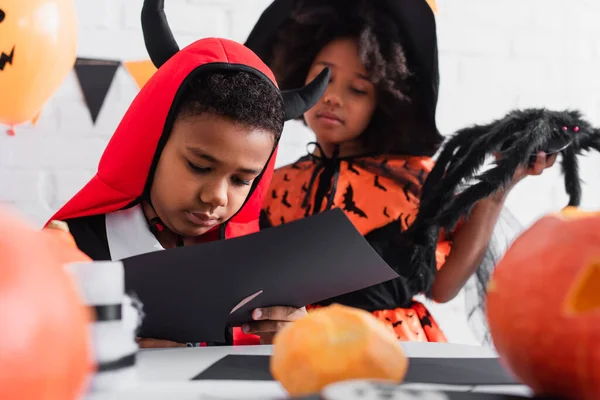 Menino afro-americano cortando papel preto perto da irmã em traje de bruxa halloween — Fotografia de Stock