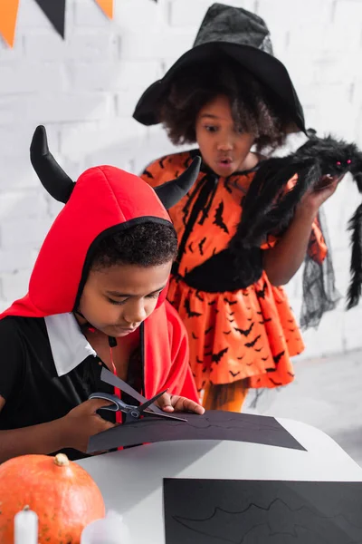 Afro-américain garçon coupe papier noir près soeur surprise en costume d'Halloween sorcière tenant araignée jouet — Photo de stock