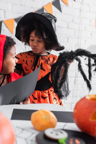 Afro-américain garçon coupe papier noir près de soeur en costume de sorcière tenant jouet araignée — Photo de stock