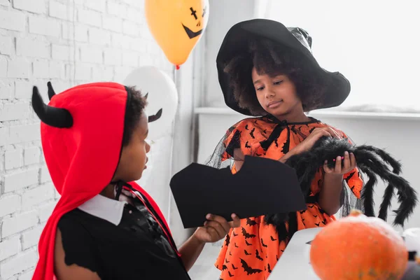 Africano americano ragazzo tenendo nero cartone vicino sorella in strega costume tenendo giocattolo ragno — Foto stock
