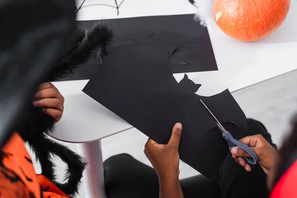 Vista recortada del niño afroamericano cortando cartón negro con plantilla de murciélago cerca de hermana borrosa sosteniendo araña de juguete - foto de stock