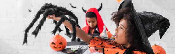 African american girl in witch costume holding toy spider near blurred brother, banner — Stock Photo
