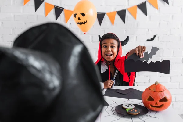 Emocionado afroamericano chico en traje de halloween mostrando plantilla de murciélago a hermana borrosa en sombrero puntiagudo - foto de stock