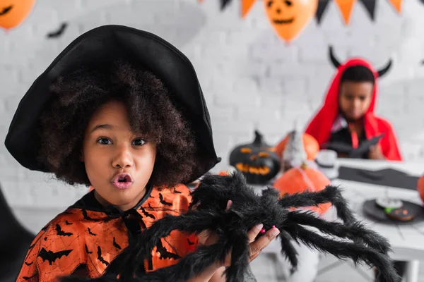 Assustador Africano americano menina em bruxa traje segurando brinquedo aranha perto borrado irmão — Fotografia de Stock