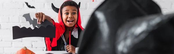 Emocionado afroamericano chico en traje de halloween mostrando plantilla de murciélago a hermana borrosa en sombrero puntiagudo, bandera - foto de stock